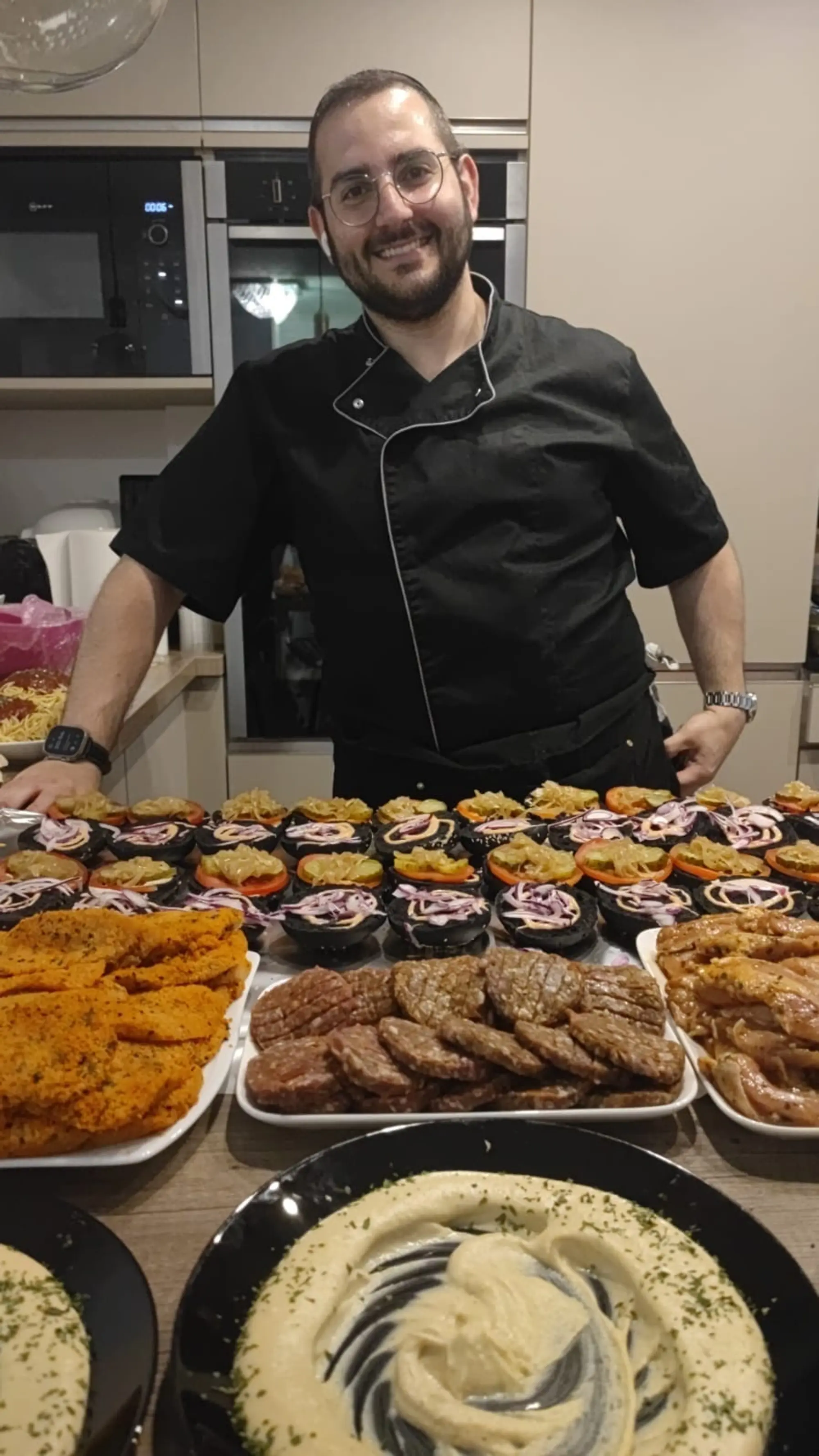 Benji souriant, en veste de chef, derrière un buffet de burgers, schnitzels et houmous
