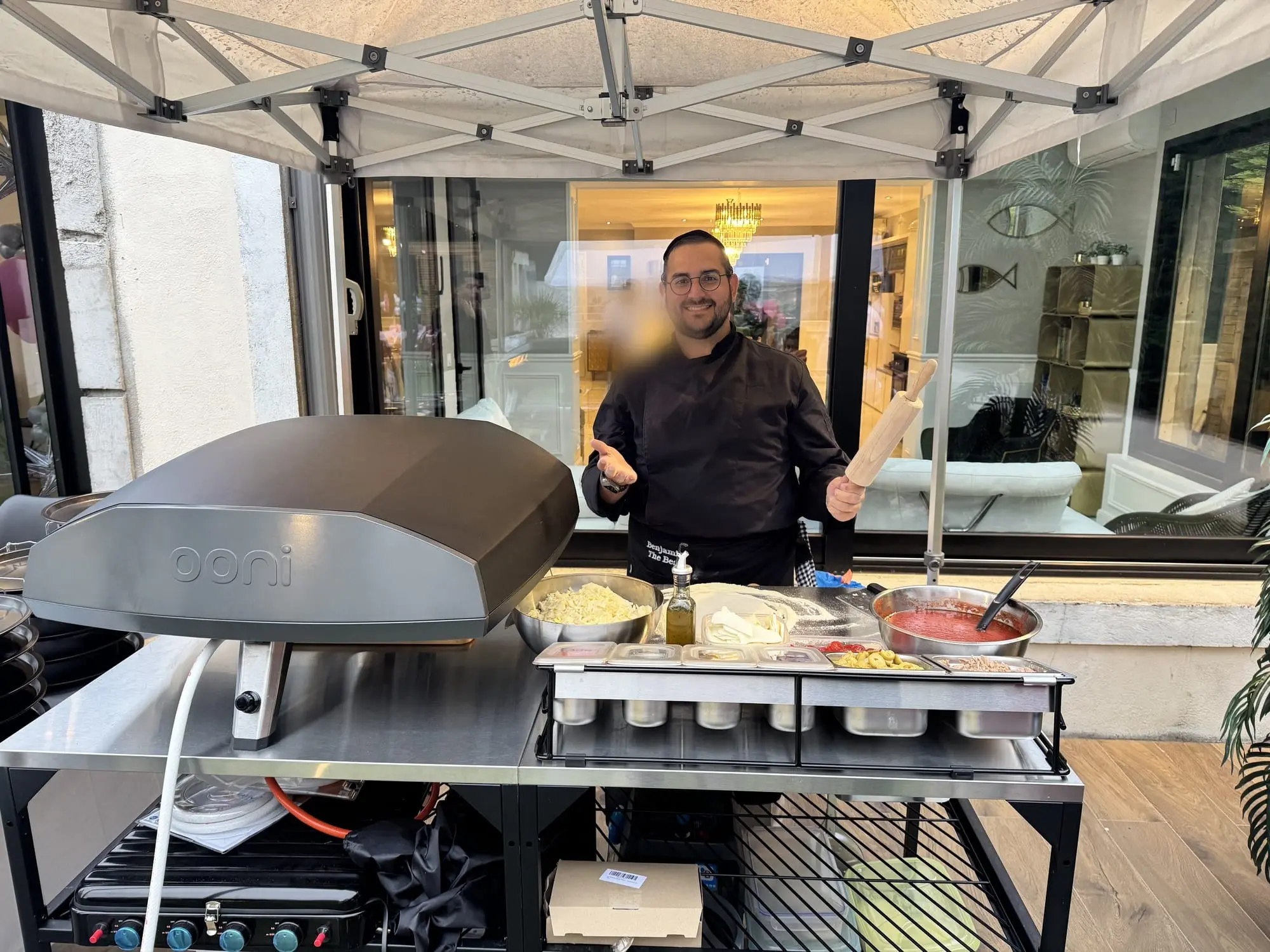 Benji en veste de chef, rouleau à pâtisserie à la main, derrière son four à pizza et ses bacs d’ingrédients, sous un barnum