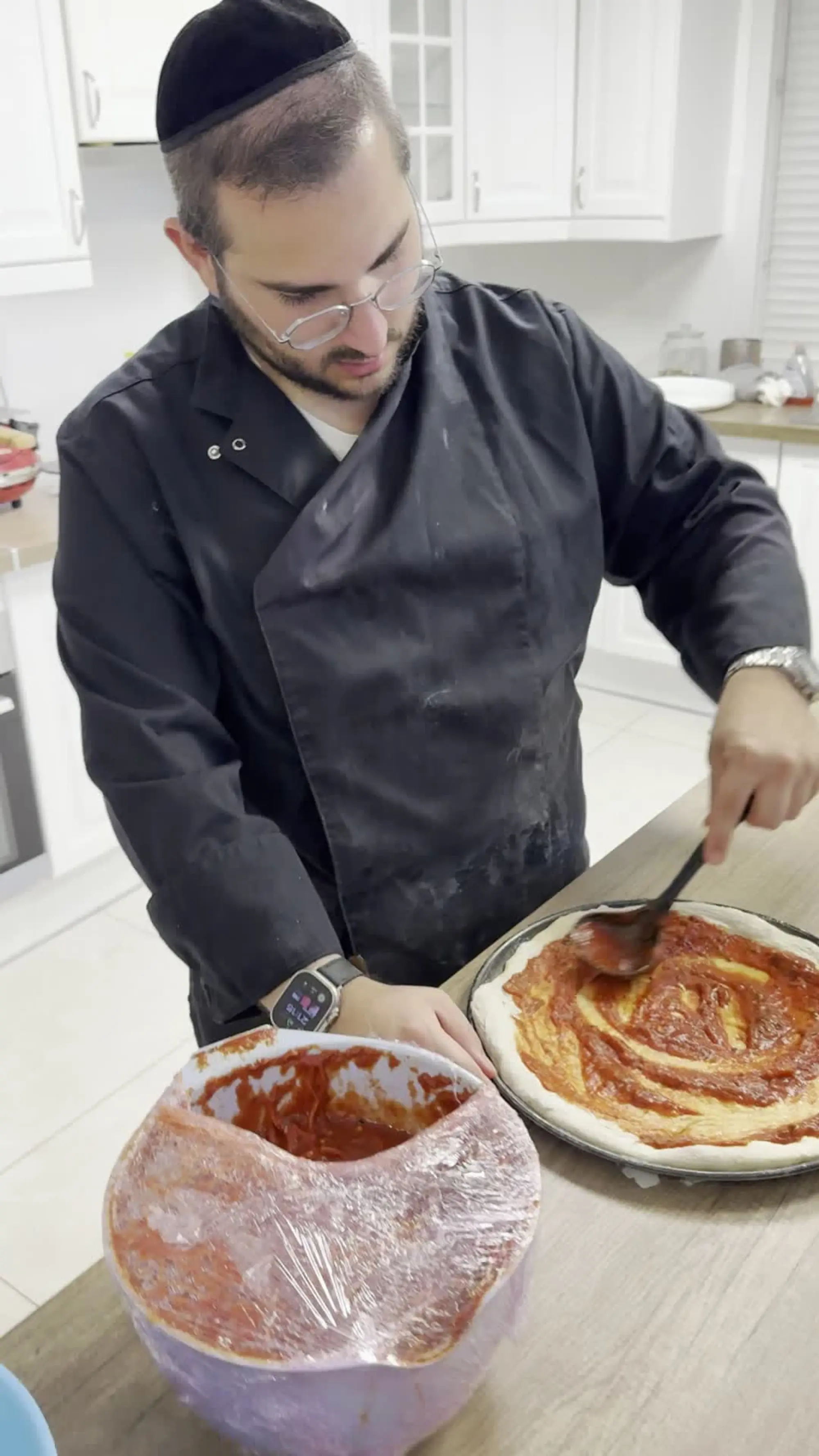 Benji en veste de chef, concentré, étale la sauce tomate à la cuillère sur une pâte à pizza