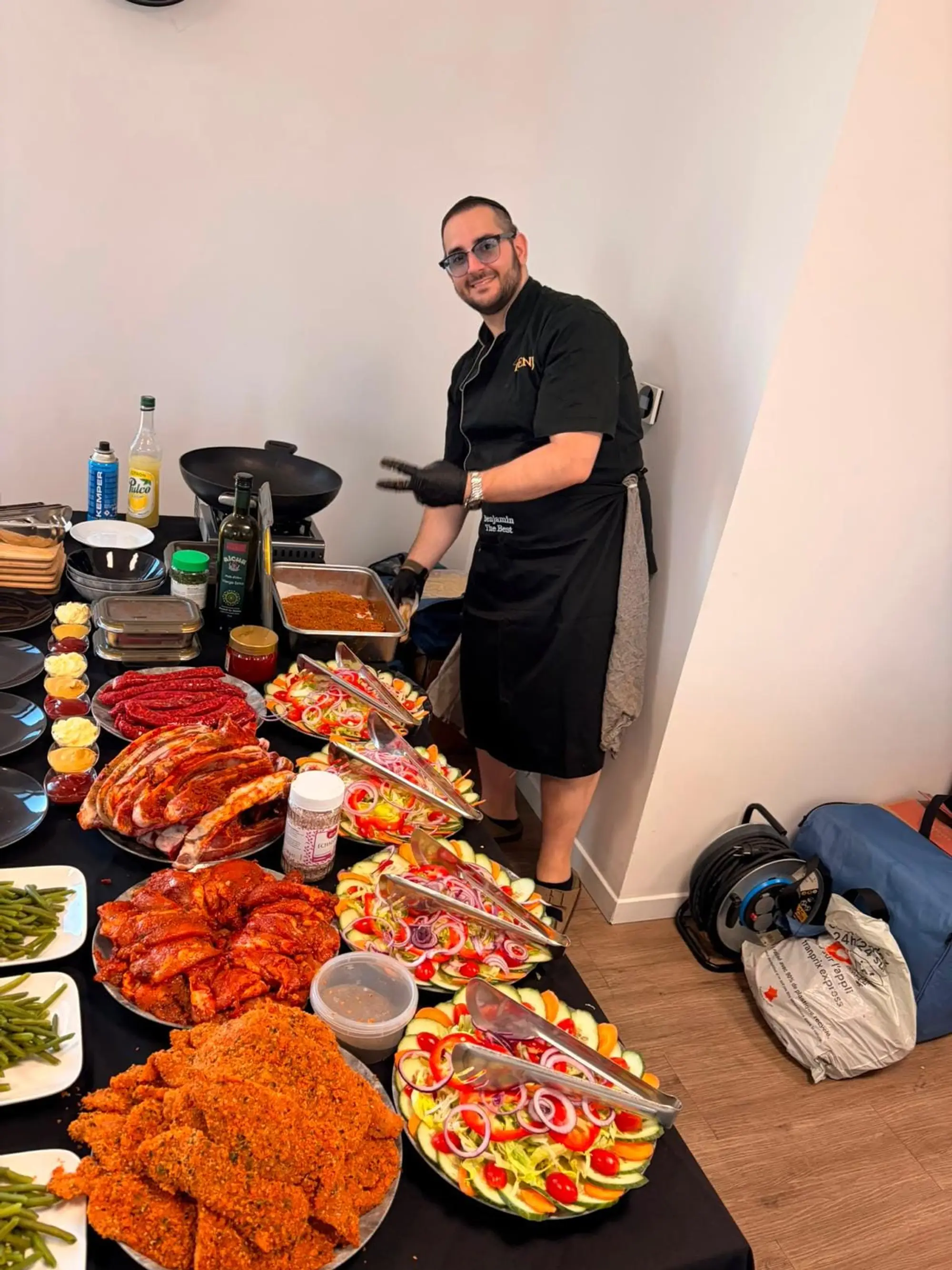Benji, en veste de chef noire et gants, sourit derrière un buffet de salades, viandes marinées et schnitzels