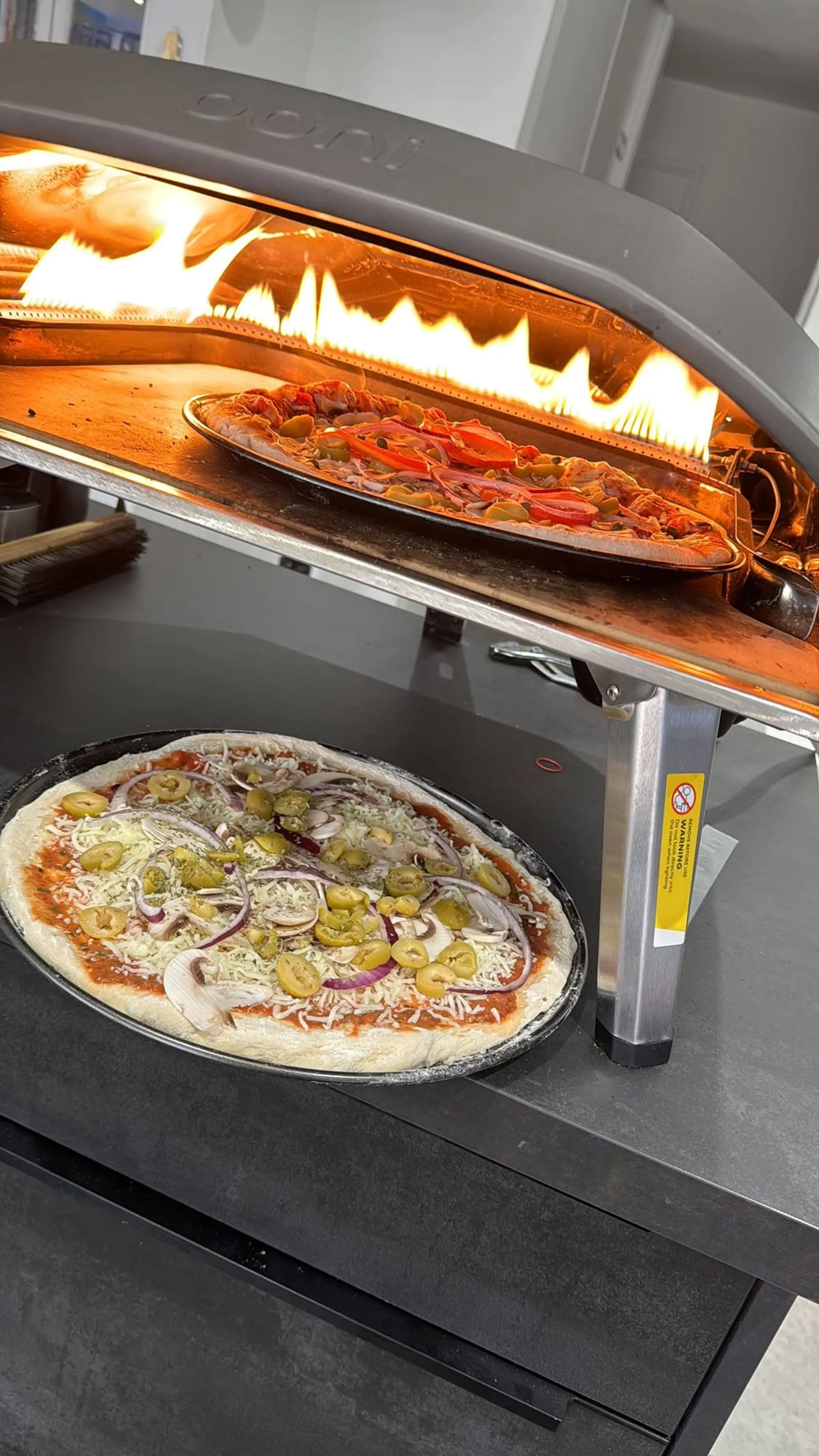 Pizza cuisant dans un four à flamme portable, une seconde pizza attend sur le plan de travail